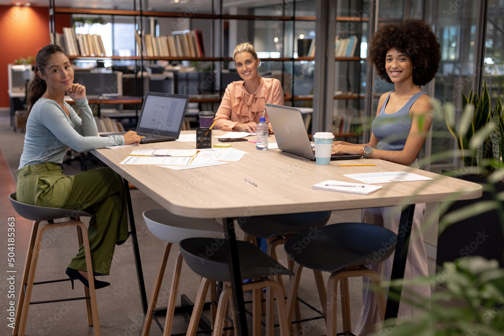 Portrait of happy multiracial female advisors sitting at desk in modern workplace