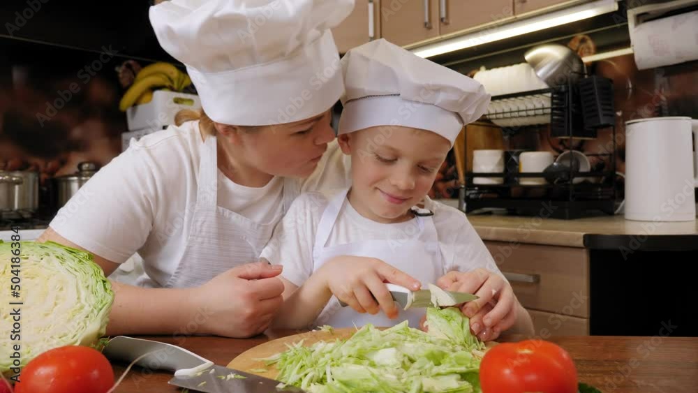 Happy curious boy learns to cut vegetables at home in the kitchen, he and his mother wear the same aprons and caps. Happy curious boy learns to cut vegetables at home in the kitchen, he and his mother wear the same aprons and caps.
