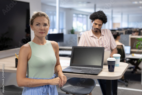 Portrait of confident cauasian businesswoman standing by laptop against biracial businesswoman