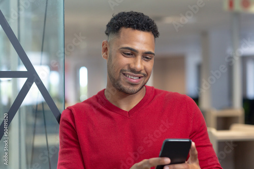 Smiling hispanic male advisor text messaging through smartphone leaning on glass wall in boardroom © WavebreakMediaMicro