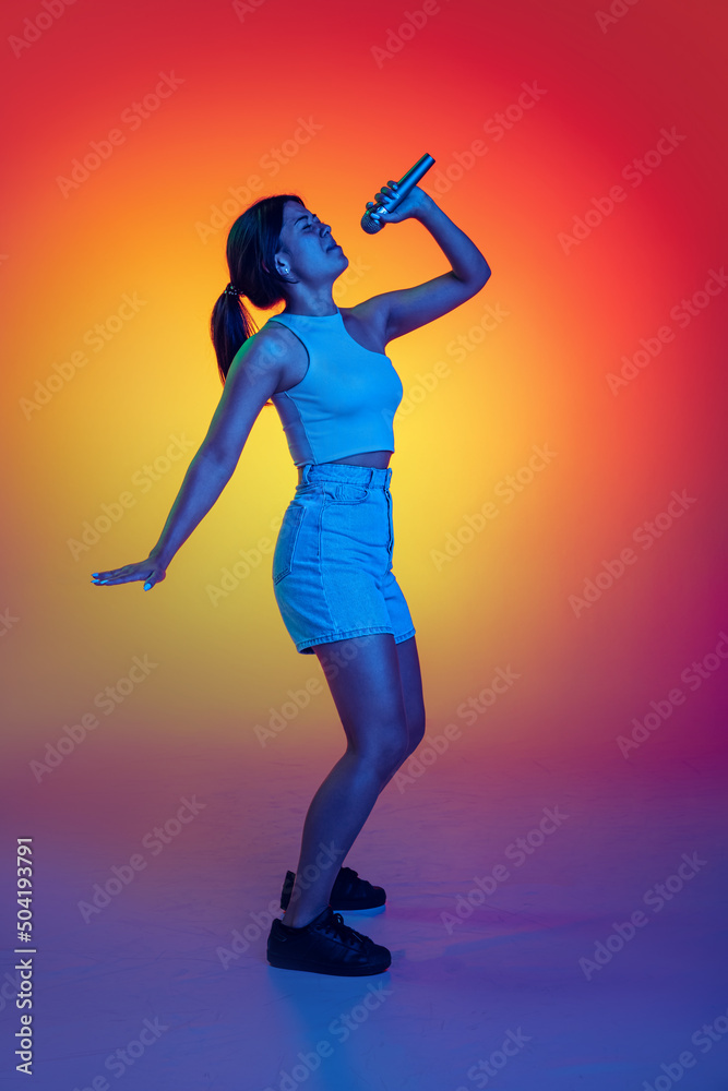 Emotive young girl singing in microphone, performing isolated over multicolored background in neon light