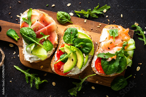 Open sandwich set with cream cheese, prosciutto, salmon, avocado and fresh greens. Top view at dark.