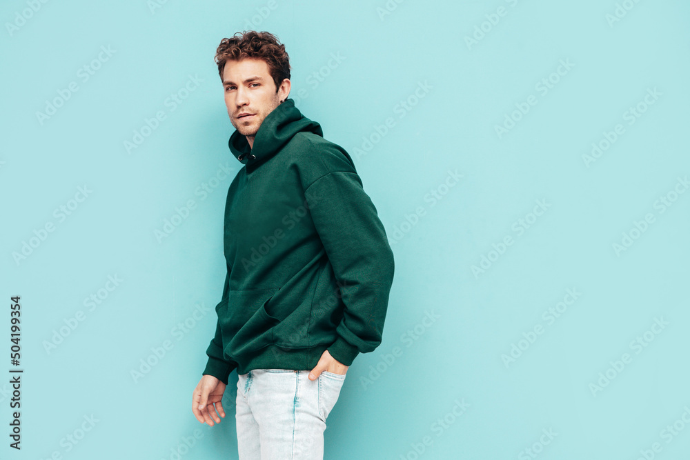 Naklejka premium Handsome confident hipster model.Sexy unshaven man dressed in summer stylish green hoodie and jeans clothes. Fashion male with curly hairstyle posing in studio. Isolated on blue