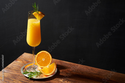 Mimosa cocktail in flute glass with orange slice and rosemary twig on a wooden bar and dark background.