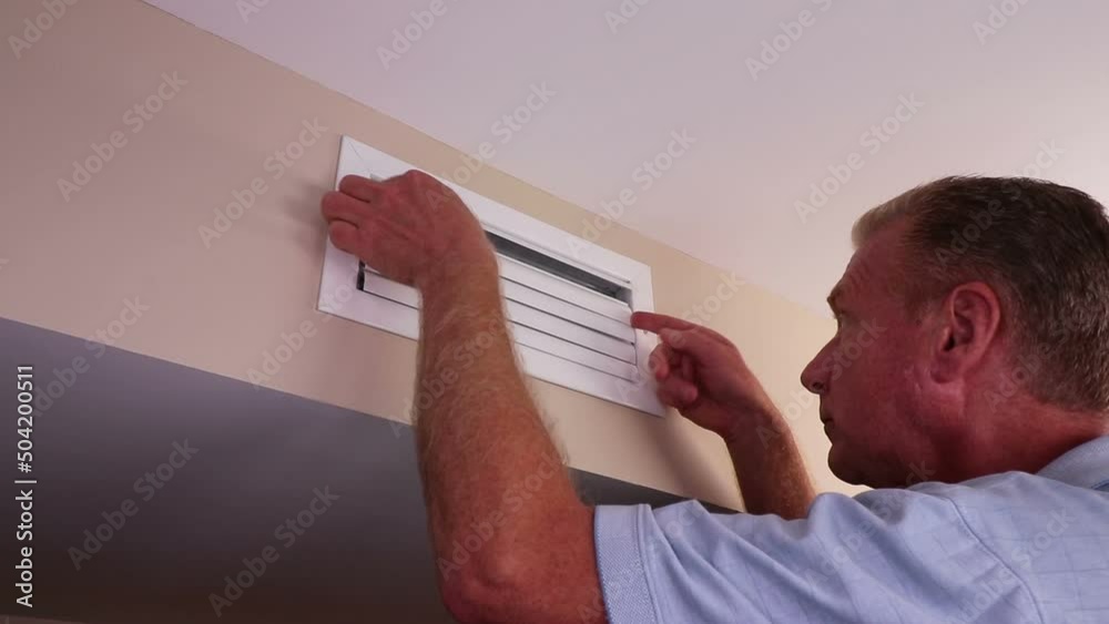 With two hands a mature adult is seen opening a home air vents adjustable register blades on a wall near a ceiling. Adjustable air vent register blades on a wall opened one at a time with both hands.
