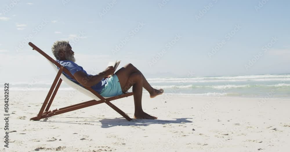 Senior african american man reading and lying on sunbed on sunny beach
