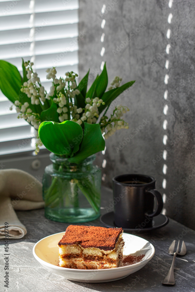 Traditional Italian dessert tiramisu, coffee and a bouquet of lilies of the valley on a gray concrete background.