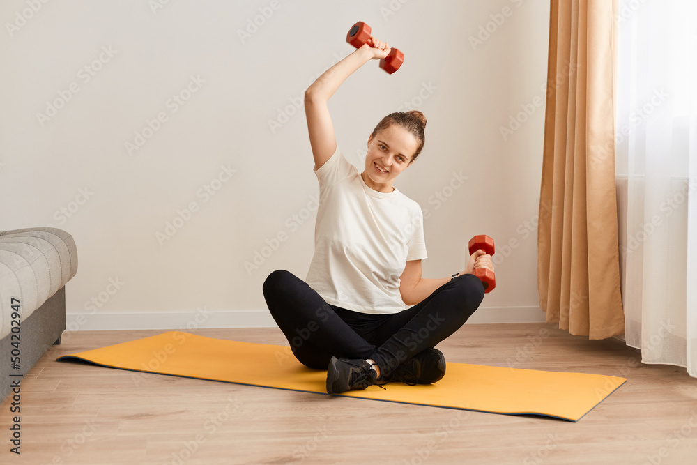 Portrait of female wearing white t shirt and black leggins exercising with dumbbells at home, training biceps and triceps, fitness, workout, healthy living and diet. Portrait of female wearing white t shirt and black leggins exercising with dumbbells at home, training biceps and triceps, fitness, workout, healthy living and diet.