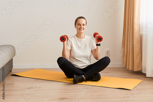 Indoor shot of smiling woman exercising with dumbbells at home, sporty beautiful young girl training at home to stay fit, female having work out in living room.
