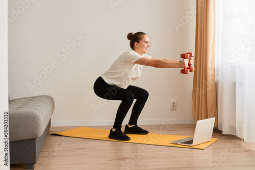 Side view of attractive woman making deep squats with dumbbells in hands in living room while watching fitness online lesson on laptop, wearing white t shirt and leggins.