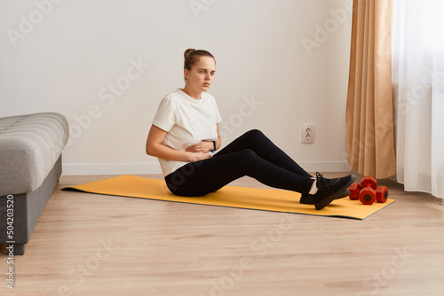 Side view portrait of unhealthy ill woman wearing white t shirt and black leggins doing sport exercises at home, sitting on yoga mat and touching painful belly, feels bad.