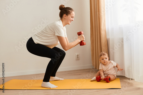 Side view portrait of slim athletic woman wearing white t shirt, standing on yoga mat, squatting with dumbbells in hands, training legs, butts and arms while looking after baby.
