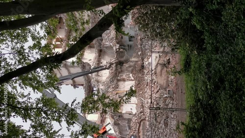 Excavator (bulldozer) demolishing a concrete building on a demolition site with debris piles. Vertical 4k still shot looking through tree branches.