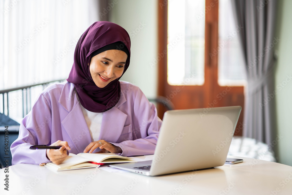 Muslim woman wearing hijab sitting at a table in a cafe working at a laptop holding a notebook with records.
