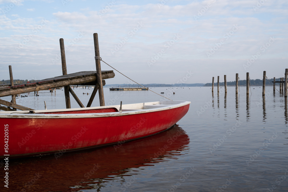Red and white rowing boat roped up at the wooden pier on a morning with a cloudy sky