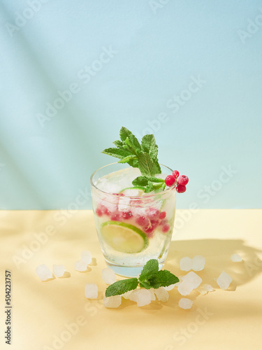 Non-alcoholic soda water tonic. Cold drink in crystal glass with ice, lime slice, redcurrant and mint lefs on yellow and blue background. photo