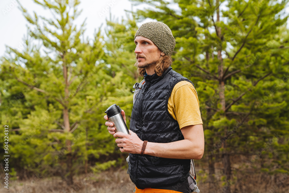 Fototapeta premium A man holds a thermos with coffee in his hands, a tourist guy is dressed in a black vest, a green hat on his head, a hike in the forest to nature, long hair hipster with a beard