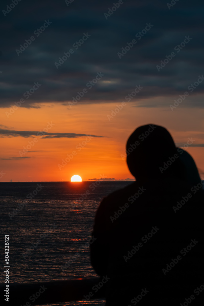 Rear view of the silhouette of a couple in love standing on the pier and enjoying the stunning sunset of the setting sun over the horizon of the sea. Vertical photo