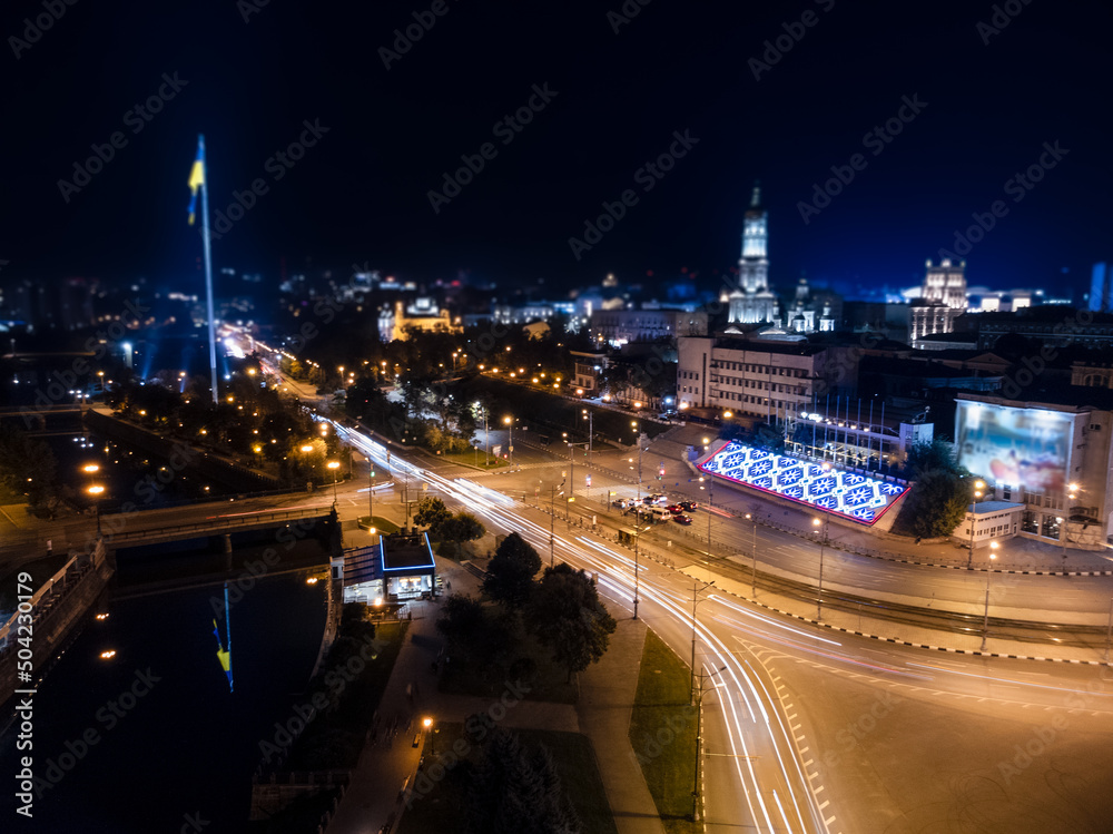 Flag of Ukraine on illuminated river embankment with reflection at night. City aerial view near Dormition Cathedral in Kharkiv, Ukraine with long exposure and background blur Flag of Ukraine on illuminated river embankment with reflection at night. City aerial view near Dormition Cathedral in Kharkiv, Ukraine with long exposure and background blur