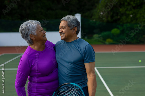 Smiling biracial senior couple embracing and looking at each other while standing in tennis court