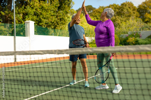 Cheerful biracial senior couple holding rackets giving high-five in tennis court against trees