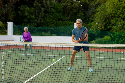 Full length of biracial senior man playing tennis with senior woman in tennis court against trees