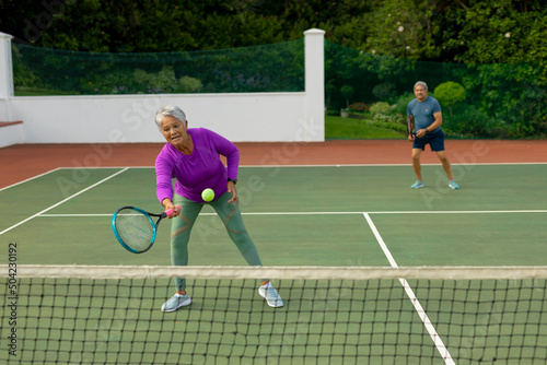 Biracial active senior woman with senior man playing tennis in tennis court against trees