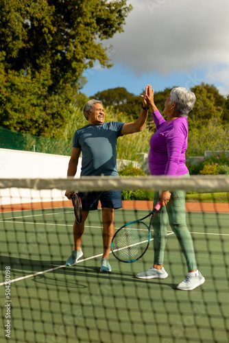 Happy biracial senior couple holding rackets giving high-five in tennis court against cloudy sky