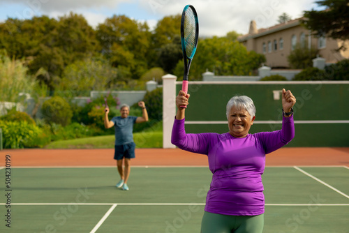 Happy biracial senior woman holding racket and gesturing at tennis court with man in background © wavebreak3