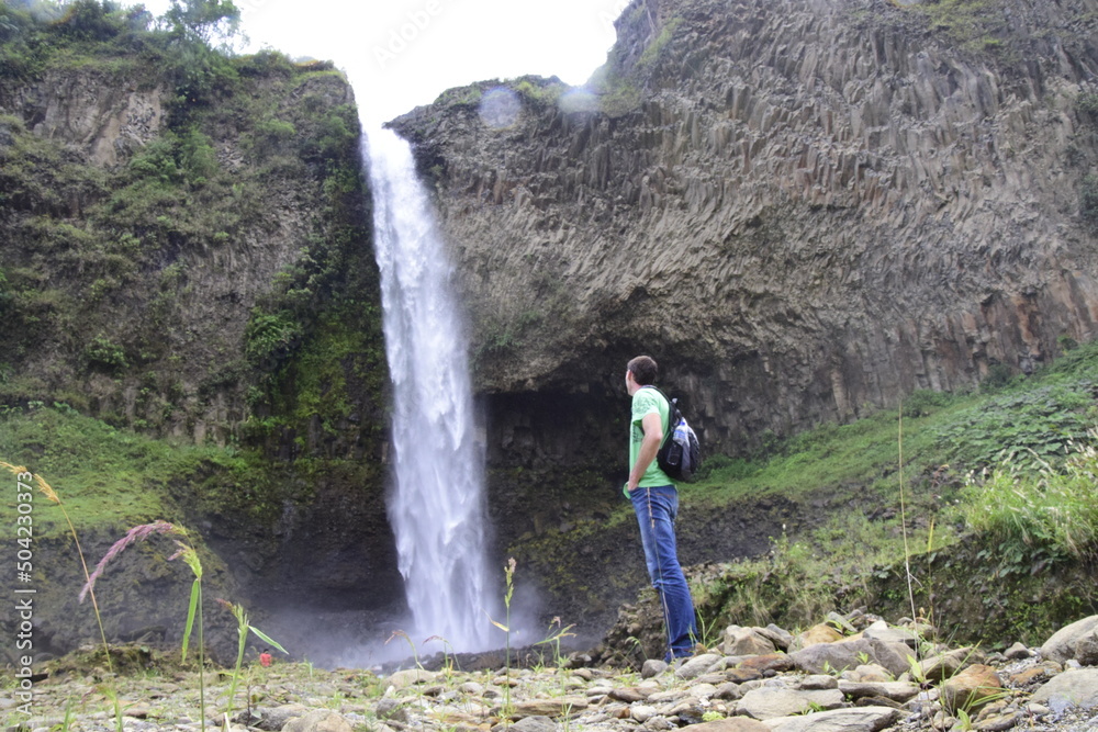 A man with a backpack is standing in front of a waterfall in Banos de Agua Santa, Cascada Manto de la Novia. Banos A man with a backpack is standing in front of a waterfall in Banos de Agua Santa, Cascada Manto de la Novia. Banos