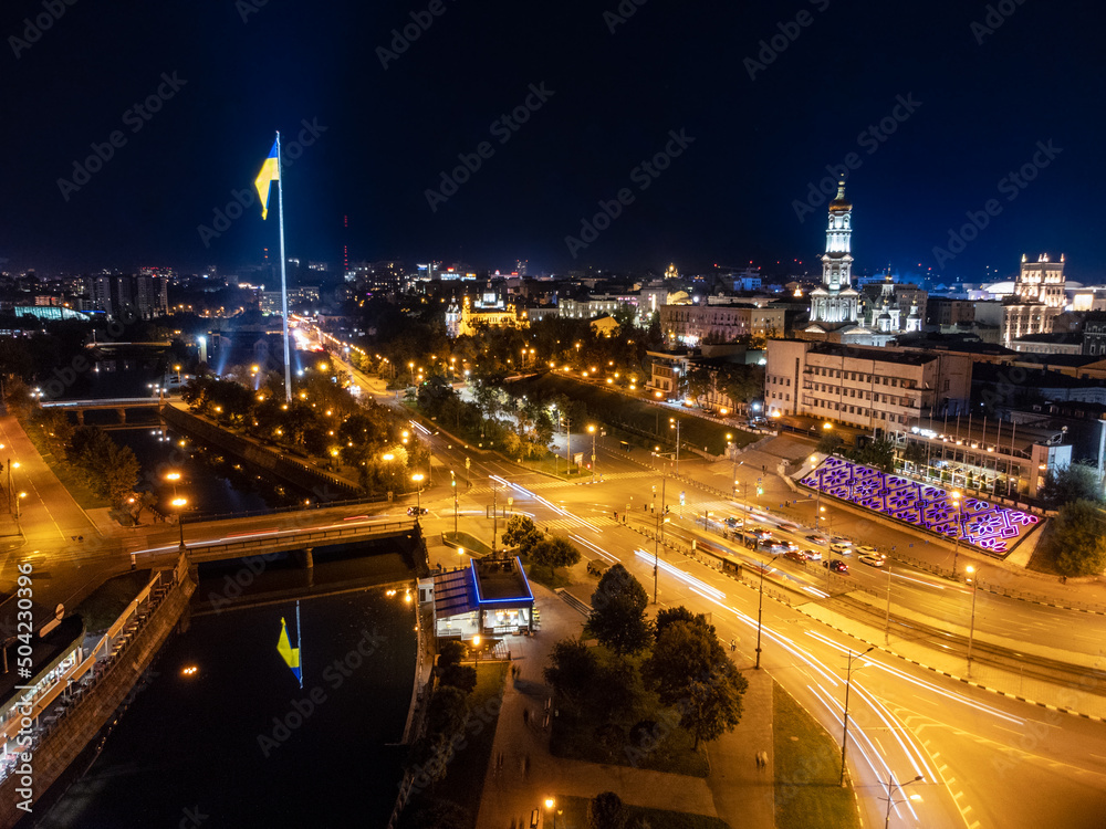 Fototapeta premium Flag of Ukraine on illuminated river embankment with reflection at night. City aerial view above river Lopan near Dormition Cathedral in Kharkiv, Ukraine with long exposure
