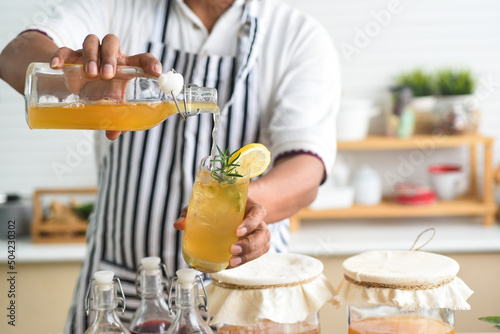 Chef's hand holding and pouring a glass of Homemade fermented kombucha tea, variety of flavors in bottles and glass jars arranged mix  fruit, scoby on wooden table. Healthy natural probiotic drink.