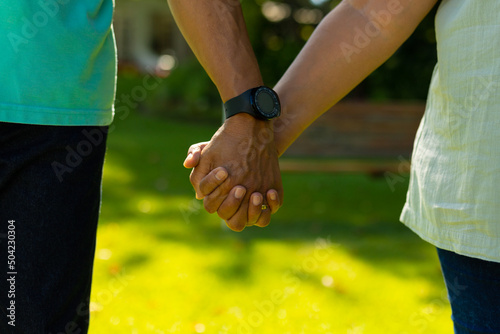 Midsection of biracial senior man wearing wristwatch holding wife's hand while standing in park