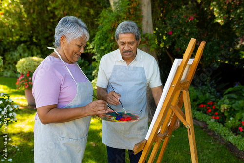 Biracial senior couple wearing aprons painting with watercolors on canvas against plants in yard