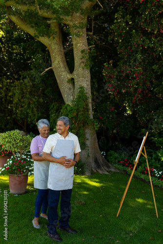 Biracial senior wife tying husband's apron standing by canvas and easel against plants in yard