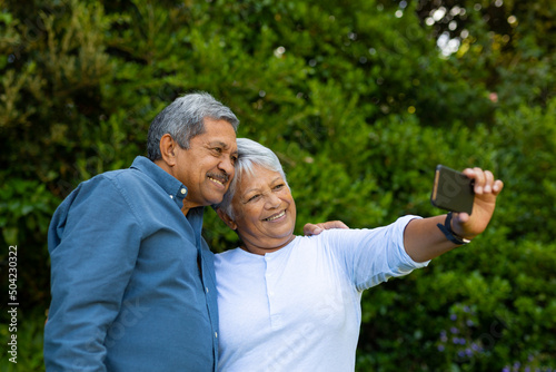 Smiling biracial senior woman taking selfie with husband over smart phone against trees in park