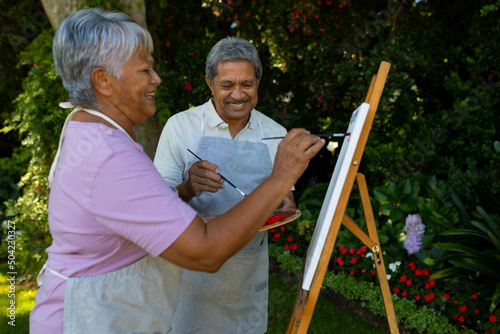 Cheerful biracial senior couple painting with watercolors on canvas against plants in yard