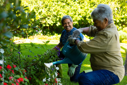 Smiling biracial senior man looking at senior wife watering flowers with watering can in yard
