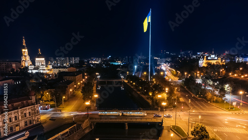 Tableau sur toile Flag of Ukraine and Holy Annunciation Cathedral on illuminated river embankment with tram on bridge across  river Lopan at night
