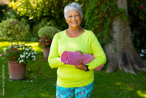 Portrait of smiling biracial senior woman wearing sports clothing holding yoga mat against plants