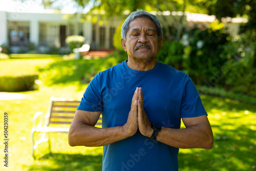 Biracial senior man with eyes closed meditating in prayer position against plants at yard