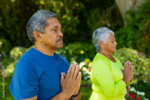 Biracial senior couple with eyes closed meditating in prayer positing while standing in yard