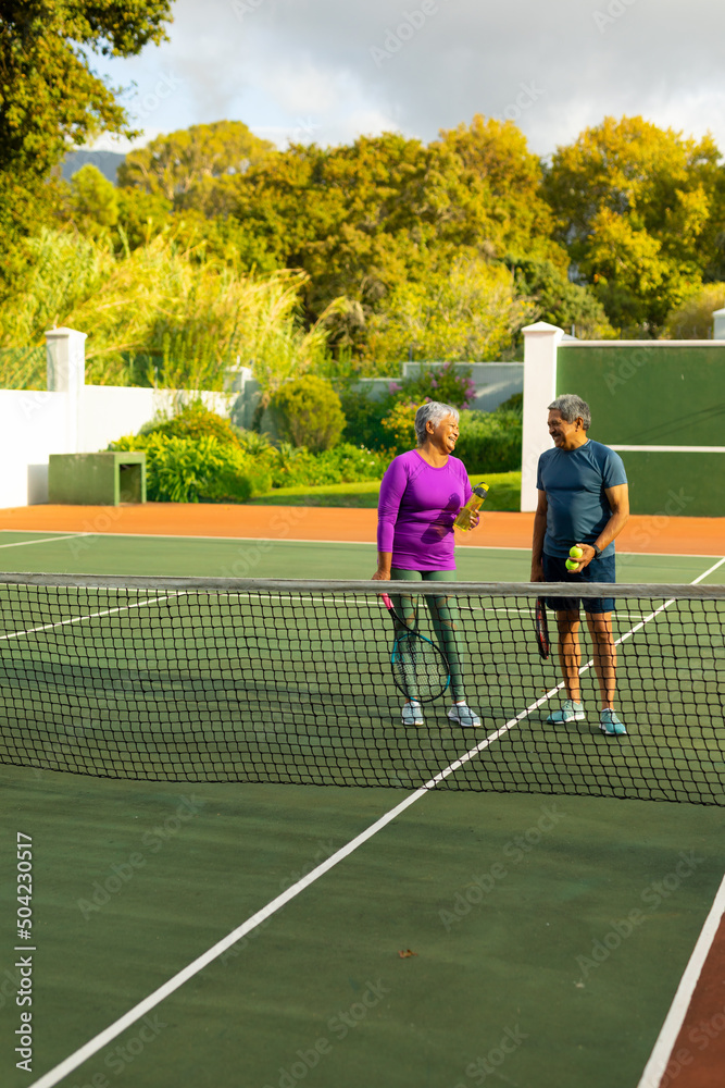Biracial senior couple holding rackets, balls and water bottle talking at tennis court against trees