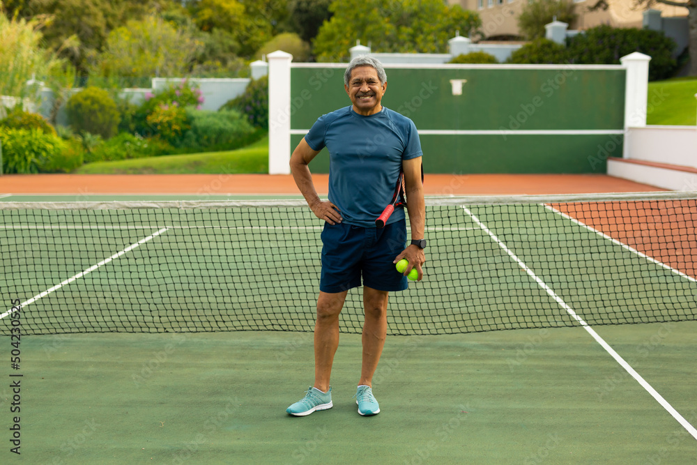 Portrait of smiling biracial senior man holding balls and racket standing by net in tennis court