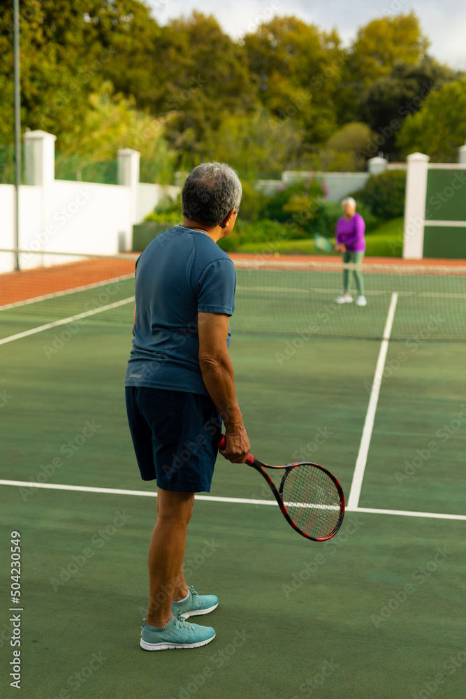 Biracial senior man holding racket playing tennis with senior wife against trees at tennis court