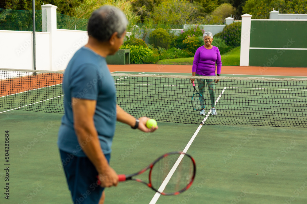 Smiling biracial senior woman playing tennis with senior husband at tennis court