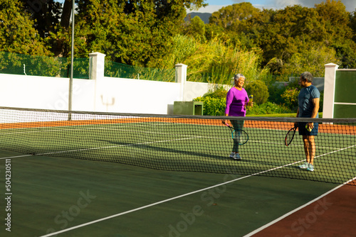 Biracial senior couple talking while standing at tennis court against trees during sunny day