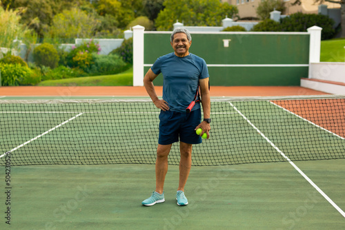 Portrait of smiling biracial senior man holding balls and racket standing by net in tennis court © WavebreakMediaMicro