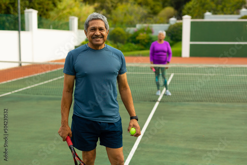 Portrait of smiling biracial senior man holding racket and ball playing tennis with wife at court