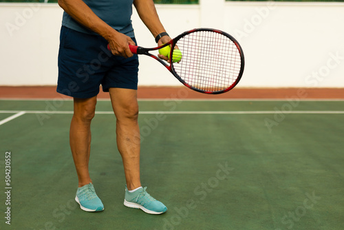 Low section of biracial senior man holding racket and ball while standing at tennis court